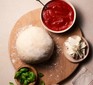 Pizza dough on a board next to a bowl of tomato sauce and a ramekin of basil